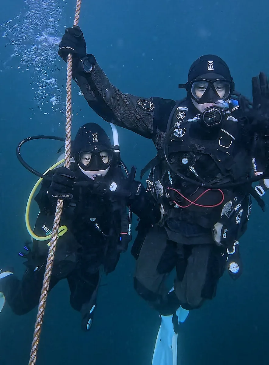 Two scuba divers descending feet-first on a line in blue water, the controlled descent that prevents scuba ear pain