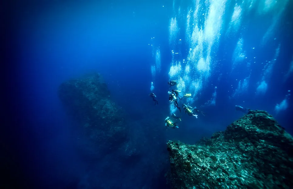 Divers descending slowly along a rocky reef in deep blue Korean water, giving their ears time to equalise